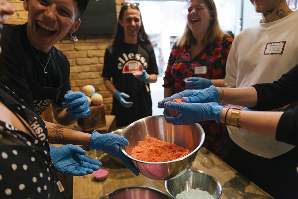 People standing around and wearing blue gloves, there is a bowl of orange bathbomb ingredients that is being held